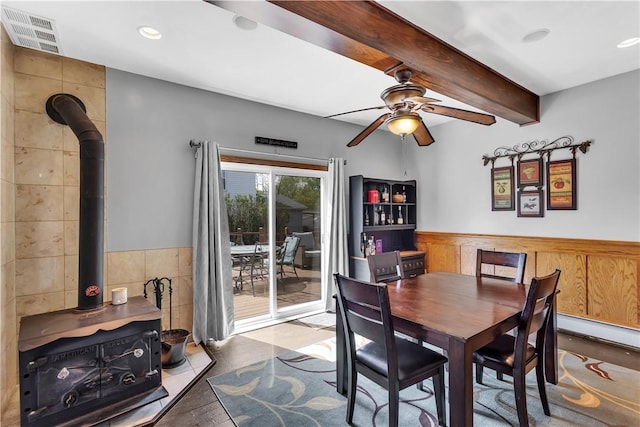 dining room featuring a wood stove, a wainscoted wall, ceiling fan, beam ceiling, and tile walls