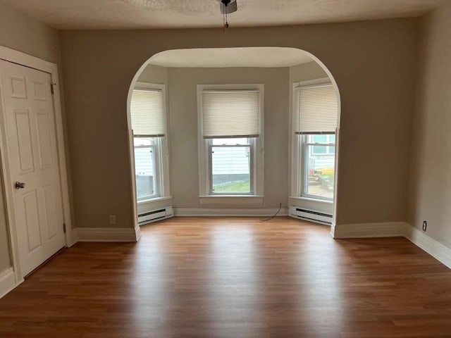 empty room featuring baseboard heating, arched walkways, wood finished floors, and baseboards