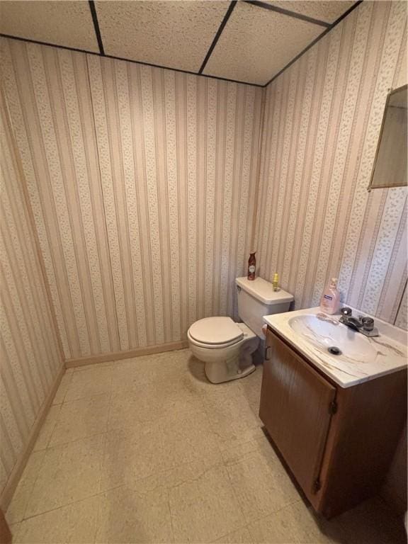 half bath with wallpapered walls, toilet, vanity, tile patterned floors, and a drop ceiling