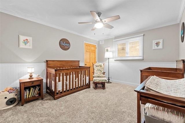 bedroom with a wainscoted wall, crown molding, carpet, a crib, and a ceiling fan
