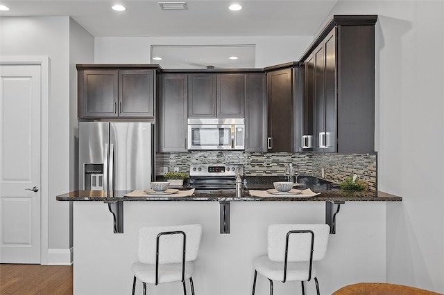 kitchen with appliances with stainless steel finishes, dark stone counters, dark brown cabinetry, decorative backsplash, and recessed lighting