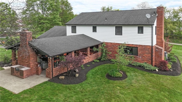 rear view of property featuring a chimney, a patio, a yard, brick siding, and a shingled roof