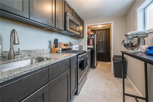 kitchen featuring appliances with stainless steel finishes, a sink, light marble finish floors, light stone counters, and dark brown cabinets