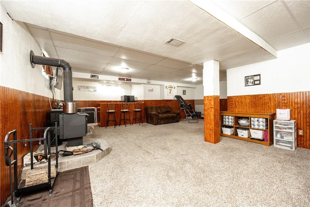 finished below grade area featuring a wood stove, wooden walls, wainscoting, carpet flooring, and a paneled ceiling