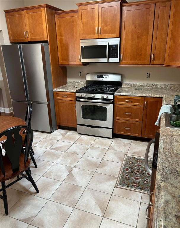 kitchen featuring appliances with stainless steel finishes, brown cabinetry, light tile patterned flooring, and light stone countertops