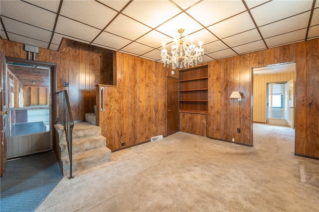 unfurnished living room featuring wood walls, a chandelier, carpet, stairway, and a paneled ceiling