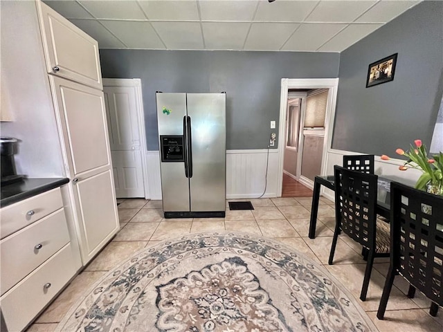 kitchen with stainless steel fridge, a drop ceiling, a wainscoted wall, light tile patterned floors, and white cabinets