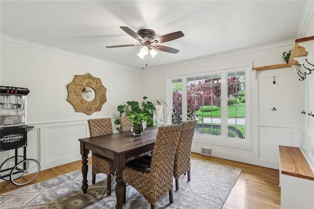 dining room featuring crown molding, ceiling fan, light wood-type flooring, a decorative wall, and a wainscoted wall