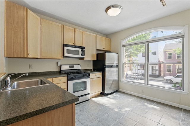 kitchen with appliances with stainless steel finishes, a sink, light brown cabinetry, dark countertops, and baseboards