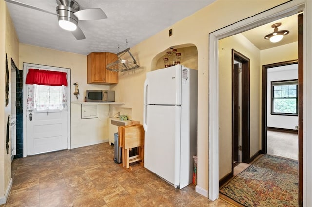 kitchen featuring freestanding refrigerator, stainless steel microwave, a ceiling fan, brown cabinetry, and baseboards