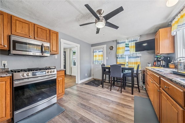 kitchen featuring stainless steel appliances, brown cabinetry, ceiling fan, light wood-style flooring, and a textured ceiling
