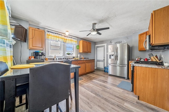 kitchen with stainless steel appliances, ceiling fan, light wood finished floors, brown cabinetry, and a textured ceiling