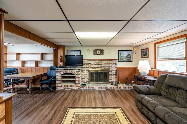 living area featuring a paneled ceiling, wood finished floors, a brick fireplace, a wainscoted wall, and wood walls