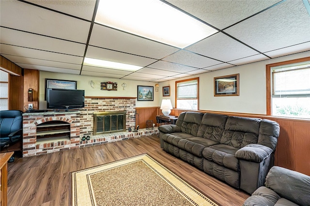 living room featuring a brick fireplace, a drop ceiling, wood finished floors, wood walls, and a wainscoted wall