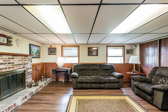 living room featuring wood finished floors, a drop ceiling, a wainscoted wall, wooden walls, and a brick fireplace