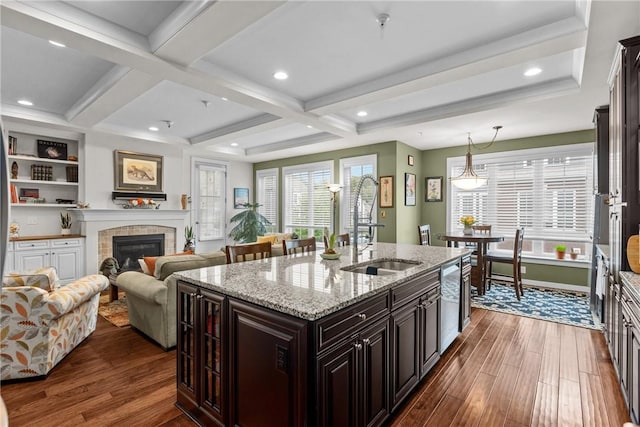 kitchen with a sink, dark wood-type flooring, a kitchen island with sink, a fireplace, and beamed ceiling