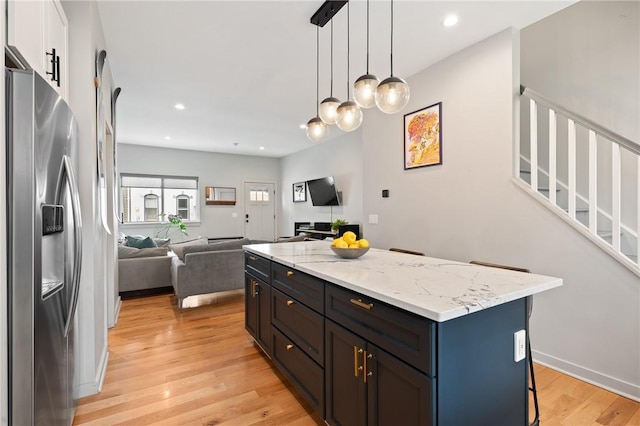 kitchen featuring stainless steel refrigerator with ice dispenser, open floor plan, light wood-style flooring, hanging light fixtures, and light stone counters