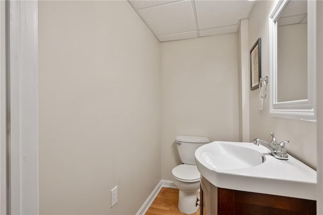 half bathroom featuring a paneled ceiling, toilet, wood finished floors, baseboards, and vanity