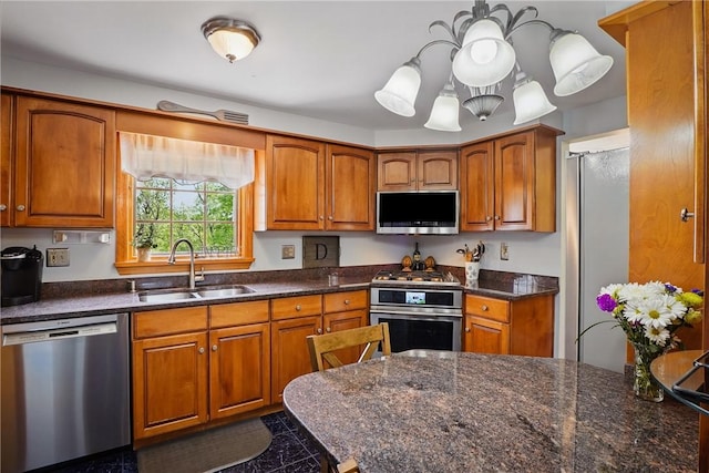 kitchen featuring stainless steel appliances, a sink, brown cabinetry, a chandelier, and dark stone counters