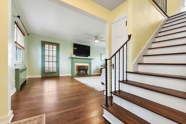 unfurnished living room with a ceiling fan, stairs, wood finished floors, a fireplace with flush hearth, and radiator