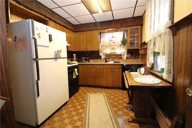 kitchen featuring freestanding refrigerator, gas stove, brown cabinets, light countertops, and a drop ceiling