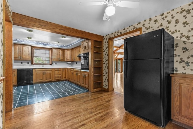 kitchen with black appliances, wallpapered walls, wood finished floors, a ceiling fan, and brown cabinetry