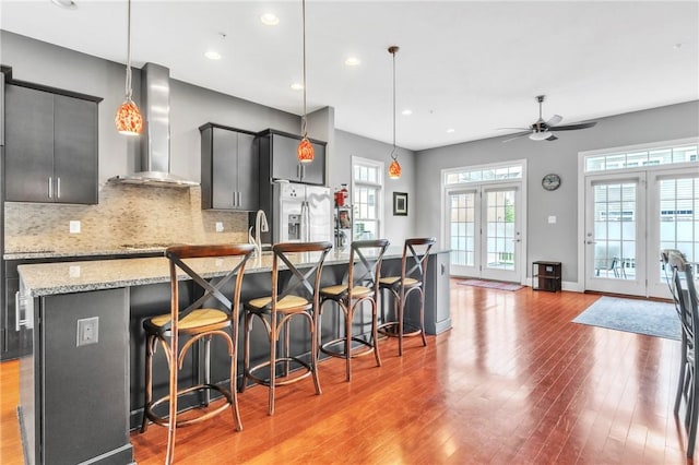 kitchen with stainless steel fridge, wall chimney exhaust hood, light wood-style flooring, a spacious island, and a ceiling fan