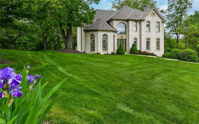 french provincial home featuring a chimney, a front lawn, roof with shingles, and brick siding