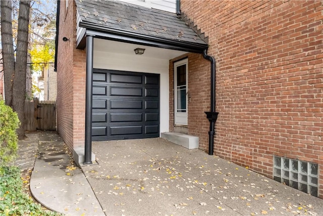 view of exterior entry with a garage, concrete driveway, brick siding, a high end roof, and fence