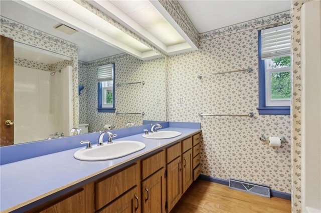 full bath featuring wood finished floors, wallpapered walls, baseboards, a shower, and double vanity