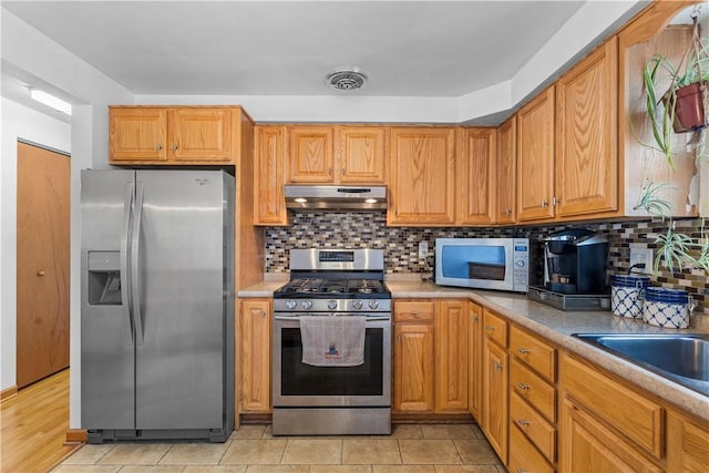 kitchen featuring appliances with stainless steel finishes, under cabinet range hood, backsplash, light countertops, and brown cabinetry