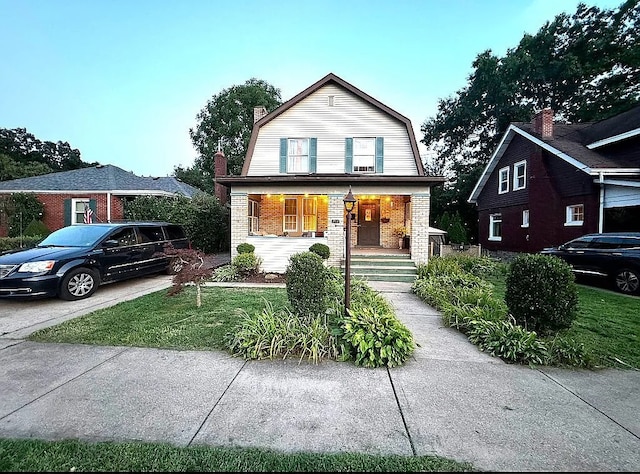 dutch colonial with a gambrel roof, covered porch, brick siding, a chimney, and a front yard
