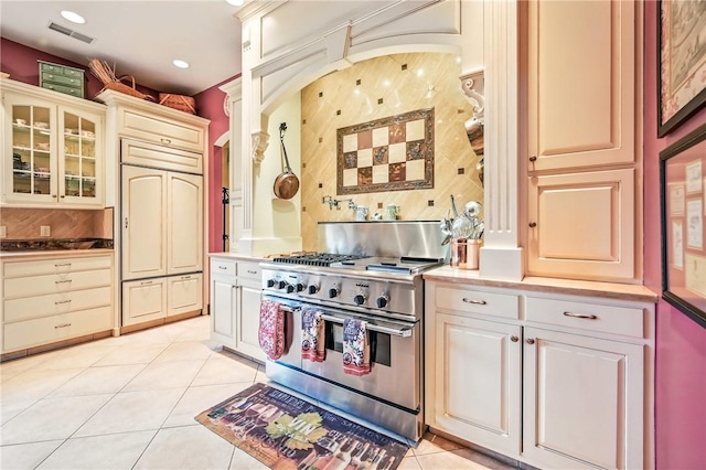 kitchen with high end stainless steel range oven, light tile patterned floors, cream cabinetry, glass insert cabinets, and light countertops