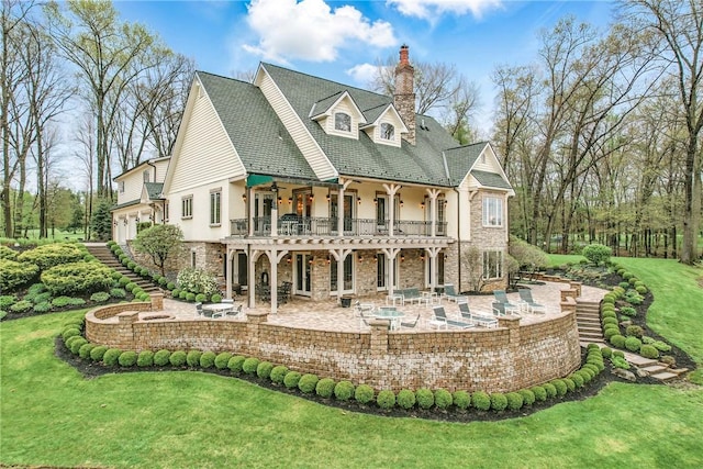 back of property with a lawn, a patio area, a chimney, stone siding, and a balcony