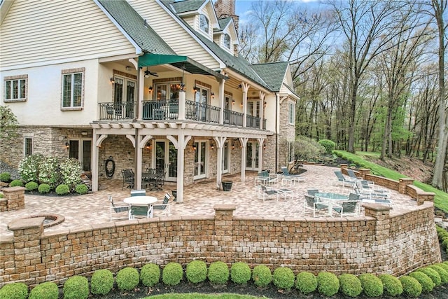 rear view of property with a balcony, ceiling fan, a patio area, french doors, and stone siding