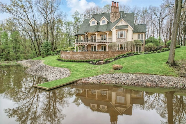 rear view of house with a chimney, a yard, stone siding, a balcony, and a water view