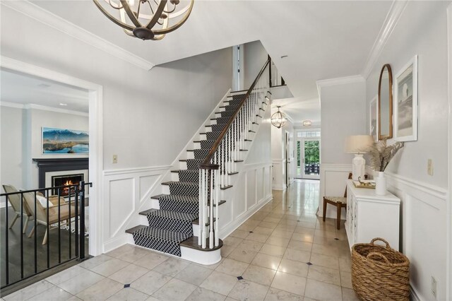 staircase featuring a chandelier, a decorative wall, ornamental molding, tile patterned flooring, and a lit fireplace