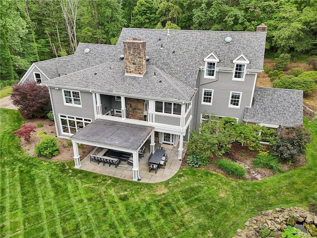 rear view of house with a chimney, a patio, a shingled roof, and a yard