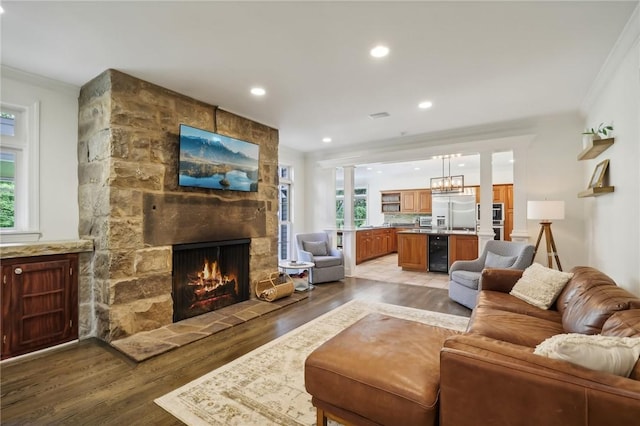 living area with beverage cooler, light wood-type flooring, a stone fireplace, ornamental molding, and healthy amount of natural light