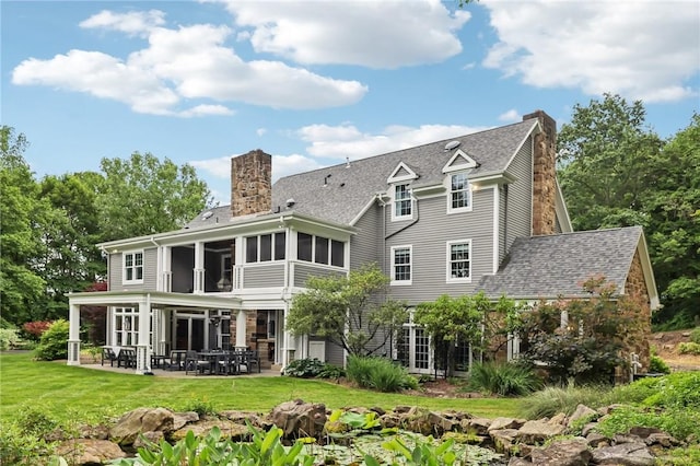 back of property featuring a chimney, a sunroom, a patio, a lawn, and roof with shingles