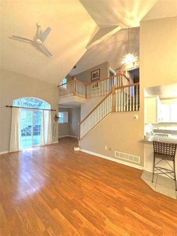 unfurnished living room with high vaulted ceiling, ceiling fan, baseboards, wood finished floors, and stairway