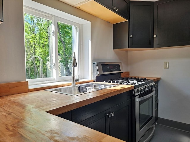 kitchen featuring stainless steel range with gas stovetop, wooden counters, dark cabinetry, a sink, and baseboards