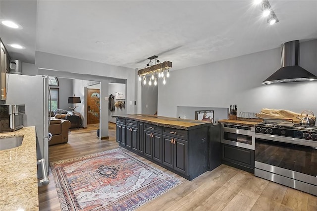 kitchen with appliances with stainless steel finishes, wall chimney exhaust hood, light wood-type flooring, wooden counters, and a sink