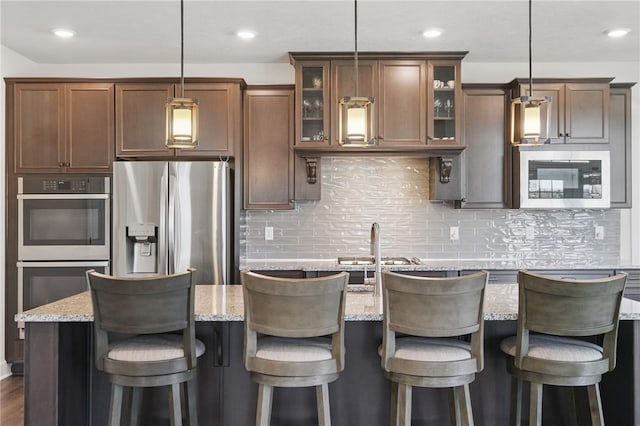5023 Kensington Dr, Cecil, PA 15317 - kitchen featuring stainless steel appliances, glass insert cabinets, a kitchen island with sink, decorative backsplash, and light stone counters