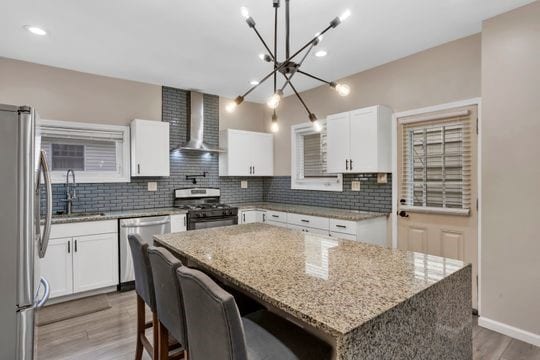 1850 Middle St, Sharpsburg, PA 15215 - kitchen featuring wall chimney range hood, appliances with stainless steel finishes, a chandelier, backsplash, and wood finished floors