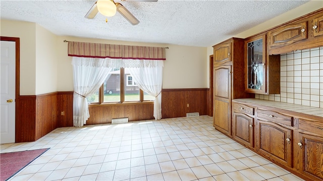 8410 Windsor Beach Ct, Harborcreek Twp, PA 16511 - kitchen featuring tile counters, a wainscoted wall, a textured ceiling, glass insert cabinets, and ceiling fan