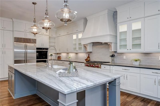 kitchen featuring appliances with stainless steel finishes, tasteful backsplash, an island with sink, custom exhaust hood, and pendant lighting