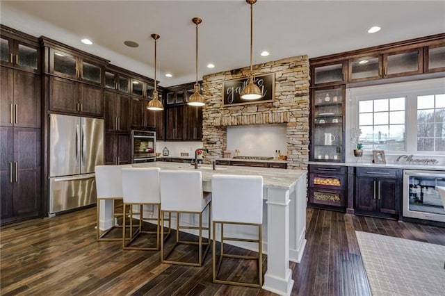 kitchen featuring dark wood-type flooring, appliances with stainless steel finishes, beverage cooler, a center island with sink, and pendant lighting