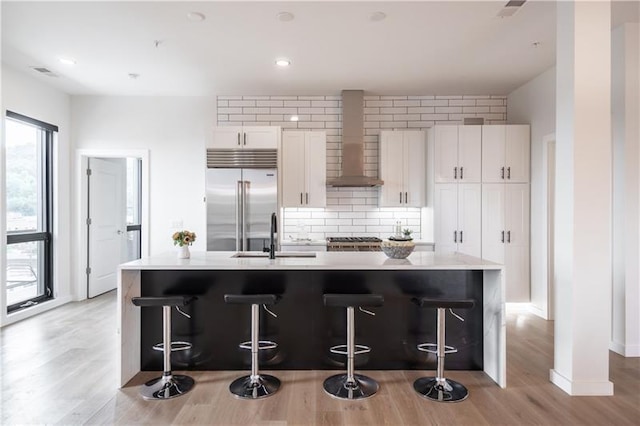 kitchen featuring wall chimney range hood, a breakfast bar, stainless steel built in refrigerator, a center island with sink, and white cabinets