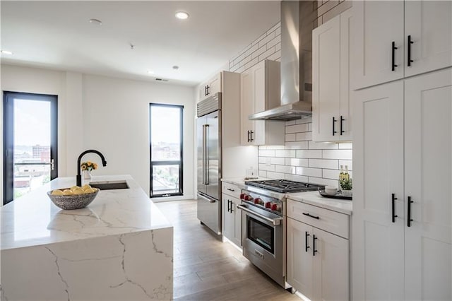 kitchen featuring sink, an island with sink, white cabinets, wall chimney range hood, and high quality appliances